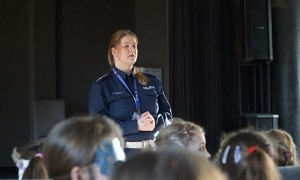 Policjantka przemawia do dzieci w sali Starogardzkiego Centrum Kultury. Na pierwszym planie krzesła i siedzące dzieci, a dalej zwrócona w ich kierunku policjantka.