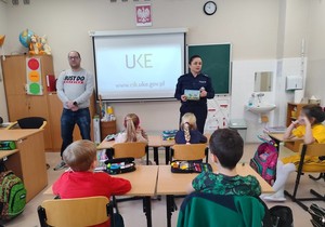Policjantka przy tablicy prowadzi zajęcia. Dzieci siedzą w ławkach szkolnych.