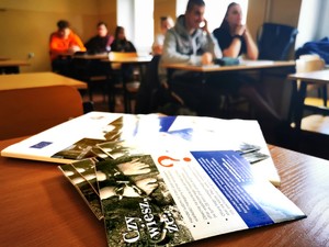 Zdjęcie w klasie. Na pierwszym planie rozłożone ulotki informacyjne. W tle siedzący w ławkach uczniowie.