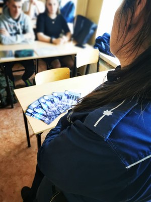 Lewe ramię policjantki zwróconej plecami zza którego widać uczniów siedzących w szkolnej ławce .