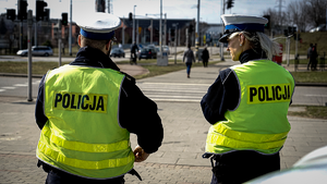 Policjanci w żółtych kamizelkach odblaskowych zwróceni plecami do aparatu obserwują pieszych korzystających z przejścia.