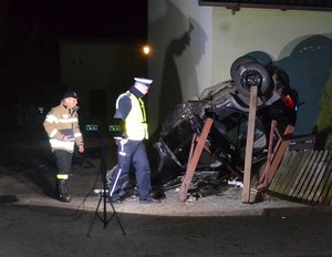Policjant prowadzi oględziny rozbitego auta. Ciemny opel leży na dachu.