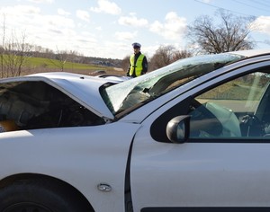 Na pierwszym planie rozbity przód peugeota, a za nim policjant z drogówki.