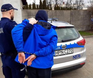 Policjant prowadzi mężczyznę, który ma założone kajdanki na rękach trzymanych z tyłu.
