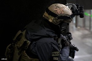 Policjant z oddziału antyterrorystycznego zwrócony tyłem patrzy przed siebie z podniesioną bronią.