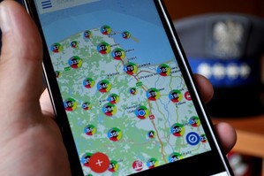aSmartfon z Krajową Mapą Zagrożeń Bezpieczeństwa, a w tle gabardynowa czapka policyjna z orzełkiem.