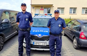Na pierwszym planie dwóch zwróconych przodem umundurowanych policjantów. Tuż za nimi radiowóz policyjny, a w tle budynek Posterunku Policji w Zblewie.