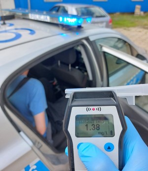 Na pierwszym planie alkotest na którego ekranie widoczne jest wskazanie, 1,38 mg/L. W tle prawy bok policyjnego radiowozu, w którym siedzi zatrzymany kierowca.