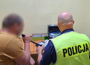 Policjant prowadzi badanie kierowcy na zawartość alkoholu w wydychanym powietrzu. Zdjęcie wykonano w budynku komendy, w pomieszczeniu, w którym znajduje się stacjonarny alkomat.