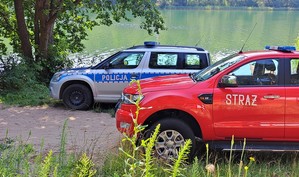 Radiowóz straży i policji. W tle szuwary, jezioro i las.