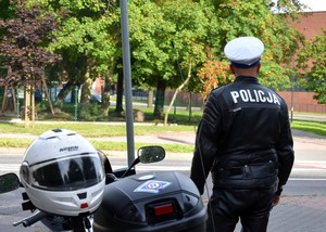 Policjant stoi przy zaparkowanym motocyklu służbowym. Na skórzanej kurtce koloru czarnego widoczny jest biały napis Policja.