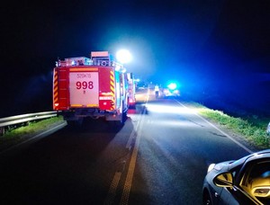 Pojazdy służb ratowniczych z włączonymi światłami błyskowymi. W oddali widoczny policjant w kamizelce odblaskowej.