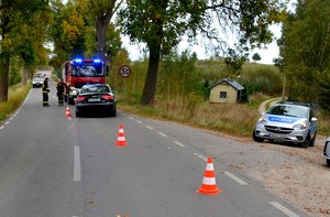 Ogólny widok na miejsce wypadku. Na jezdni stoi wóz strażacki i uszkodzone audi. W bocznej drodze zaparkowany policyjny radiowóz.