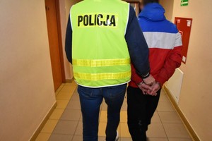 Policjant w żółtej kamizelce odblaskowej prowadzi zatrzymanego mężczyznę, który ma założone na dłoniach kajdanki.