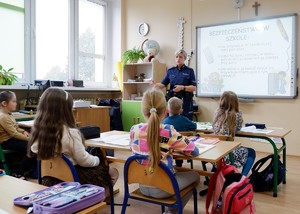 Policjantka prowadzi zajęcia z dziećmi. Dzieci siedzące w ławce patrzą na pokaz slajdów wyświetlany na tablicy. Po lewej stronie stoi dzielnicowa, która opowiada o bezpiecznej drodze do szkoły.