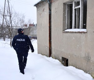 Policjant sprawdzający obejście w pobliżu pustostanu. Po prawej stronie budynek z wybitymi szybami w oknach.