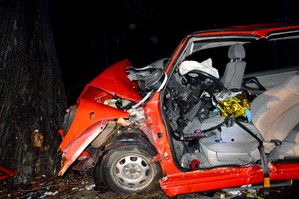 Rozbity na drzewie volkswagen. Po lewej stronie drzewo, a po prawej uszkodzone auto. Pogięta czerwona karoseria, pełno połamanych plastików.