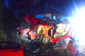 Rozbity na drzewie volkswagen. Po lewej stronie drzewo, a po prawej uszkodzone auto. Pogięta czerwona karoseria, pełno połamanych plastików.