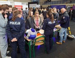 Policjanci na targach edukacyjnych EDU DAY stoją przy swoim punkcie informacyjnym i rozmawiają z maturzystami.