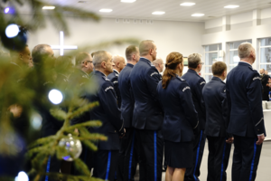 Z lewej strony choinka, a po jej prawej stronie stojący w szeregu policjanci. Choinka stoi na pierwszym planie, a policjanci znajdują się na drugim planie.