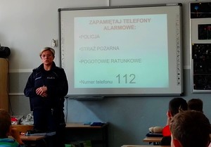 Policjantka prowadzi zajęcia z uczniami w klasie. Policjantka stoi przy tablicy, a uczniowie siedzą w ławkach.