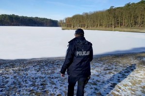 Policjantka stoi przy brzegu zamarzniętego jeziora. Na drugim planie znajdują się liczne drzewa.