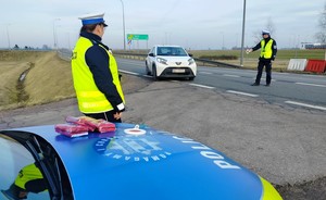 Policjanci zatrzymują do kontroli drogowej białe auto osobowe. Policjant trzyma w dłoni tarczę do zatrzymywania pojazdów.