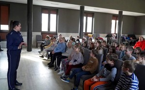 Policjantka stoi na sali i przemawia do siedzących na krzesłach dzieci.