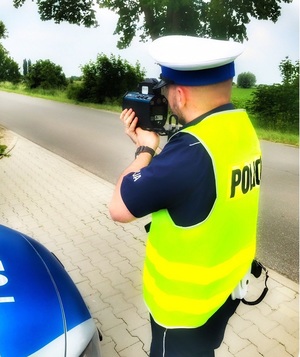 Policjant z drogówki w białej czapce i żółtej kamizelce odblaskowej mierzy prędkość radarem.