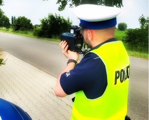 Policjant z drogówki w białej czapce i żółtej kamizelce odblaskowej mierzy prędkość radarem.