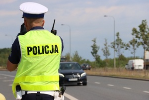 Policjant stojąc przy drodze mierzy prędkość pojazdów.