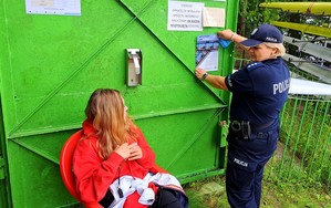 Policjantka wskazuje miejsce na drzwiach  budynku przystani, w którym można zawiesić plakat o konkursie plastycznym pn. Artystyczny przewodnik.