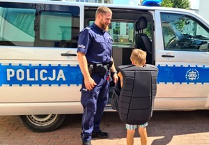 Policjant stoi z dzieckiem z boku radiowozu, który ma otwarte drzwi. Chłopiec ma założoną policyjną kamizelkę.