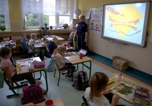 Policjantka w klasie przy tablicy opowiada dzieciom o bezpiecznej drodze do szkoły.