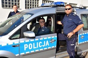 Radiowóz policyjny, za kierownicą siedzi policjanta, a przy otwartych drzwiach stoi dwóch policjantów.