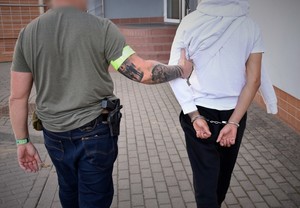 Policjant prowadzi poszukiwanego Europejskim Nakazem Aresztowania. Mężczyzna ma założone kajdanki. Policjant po cywilnemu, przy pasie widoczny pistolet, a na prawym ramieniu założona opaska odblaskowa z napisem Policja.