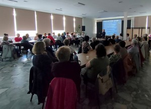 Policjantka na spotkaniu z seniorami opowiada o bezpieczeństwie. Sala ze stołami, przy których siedzą seniorzy. Ekran rzutnika z wyświetlonym slajdem.