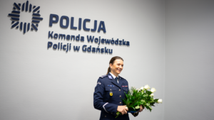 Pani Komendant trzyma wręczone na uroczystości kwiaty. W tle na białej ścianie logo i napis Policja Komenda Wojewódzka Policji w Gdańsku.