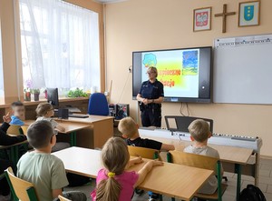Policjantka przy tablicy szkolnej prowadzi zajęcia z uczniami w klasie. Na tablicy czerwony napis o treści Bezpieczne wakacje.