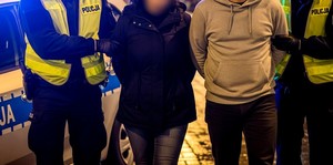 Policjanci z kobietą i mężczyzną na tle radiowozu. Zdjęcie wykadrowane w taki sposób, że nie widać twarzy osób na zdjęciu.
