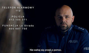 Kadr z filmu. Policjant w ciemnym pomieszczeniu, a po lewej strony biały napis z informacją o numerach alarmowych.