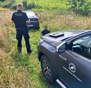 Policjant pilnuje zatrzymanego mężczyzny. Po prawej stronie pojazd służbowy nadleśnictwa.