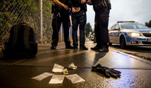 Zdjęcie ilustrujące. Na pierwszym planie woreczki z narkotykami, czarny plecak i gumowe rękawiczki. W centralnej części policjanci zakładają zatrzymanemu mężczyźnie kajdanki. Po prawej stronie stoi zaparkowany radiowóz.