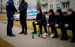Ławeczka na której siedzi kilku mężczyzn. Na chodniku przed nimi stoją butelki po alkoholu. Z lewej strony policjant legitymuje mężczyznę. W tle radiowóz z włączonym światłem błyskowym oraz bloki mieszkalne.