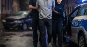 Zdjęcie ilustracyjne. Policjant z policjantką prowadzą zatrzymanego mężczyznę. W pobliżu stoi radiowóz włączonym światłem błyskowym.