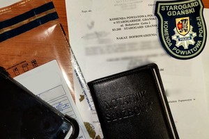 Biurko na którym leży pisemny nakaz doprowadzenia z zablurowanym tekstem, telefon komórkowy, worek foliowy z zabezpieczonym suszem roślinnym oraz naszywka naramienna z logiem Komendy Powiatowej Policji w Starogardzie Gdańskim