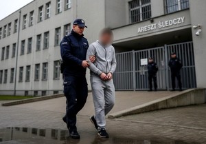 Policjant prowadzi zatrzymanego, który ma założone kajdanki. Na drugim planie budynek Aresztu Śledczego.
