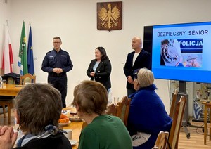 Zdjęcie organizatorów spotkania w świetlicy. W tle godło oraz ekran, na którym wyświetlana jest slajd zatytułowany: Bezpieczny senior.