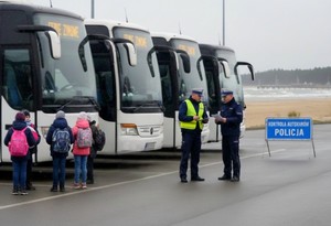 Zdjęcie obrazujące policjantów stojących przy autokarach. W pobliżu autobusów stoją dzieci z plecakami, które oczekują na wyjazd.