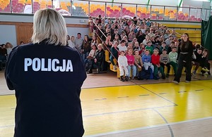 Policjantka przemawia do dzieci siedzących na ławkach w hali sportowej.